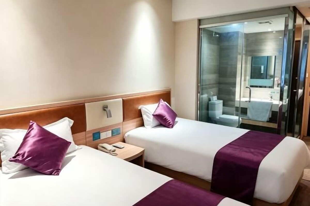 Habitación de hotel con dos camas, una mesita de noche pequeña, un baño visible a través de una puerta de cristal y una zona de ducha.