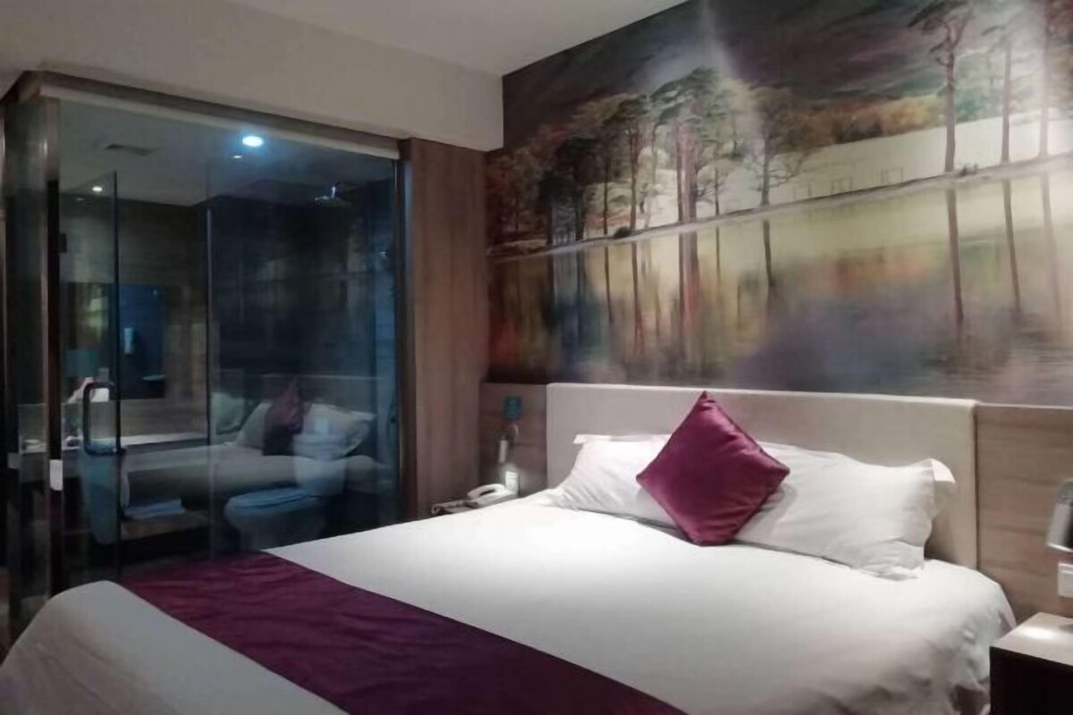 A hotel room with a large bed, a glass partition, and a painting on the wall.