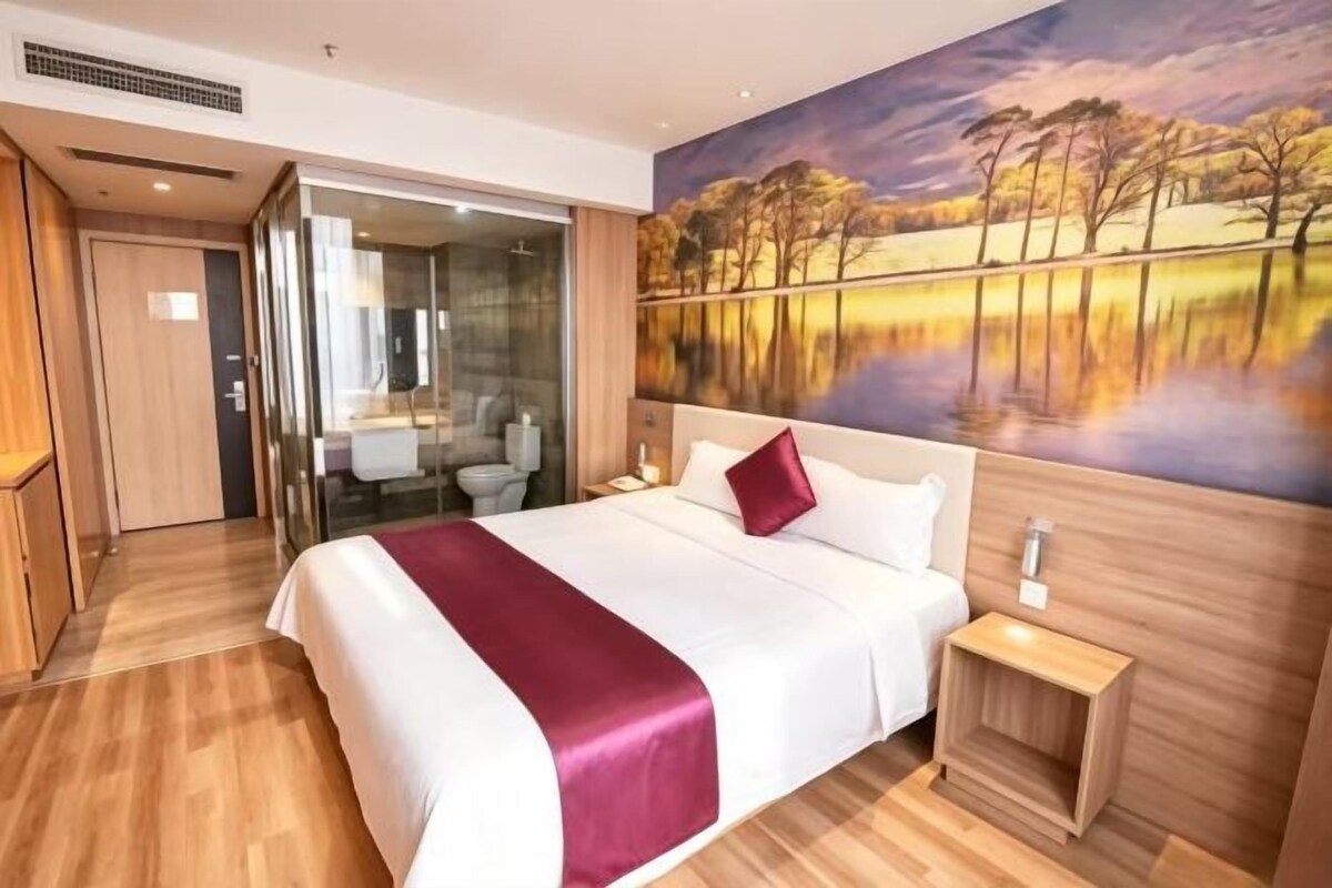 Habitación de hotel con una cama grande, un cuadro de un paisaje, piso de madera y un baño visible a través de una puerta de vidrio.