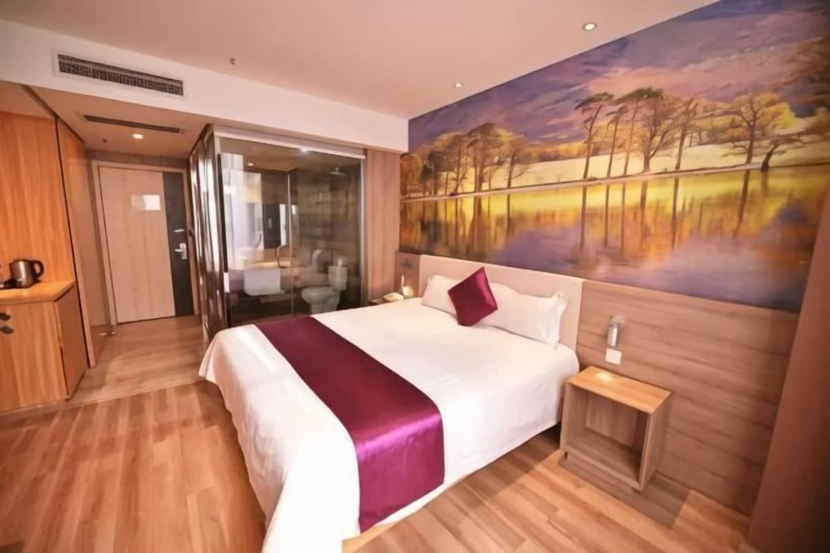 A hotel room with a large bed, a wooden floor, a painting of a landscape, and a glass door leading to a bathroom.