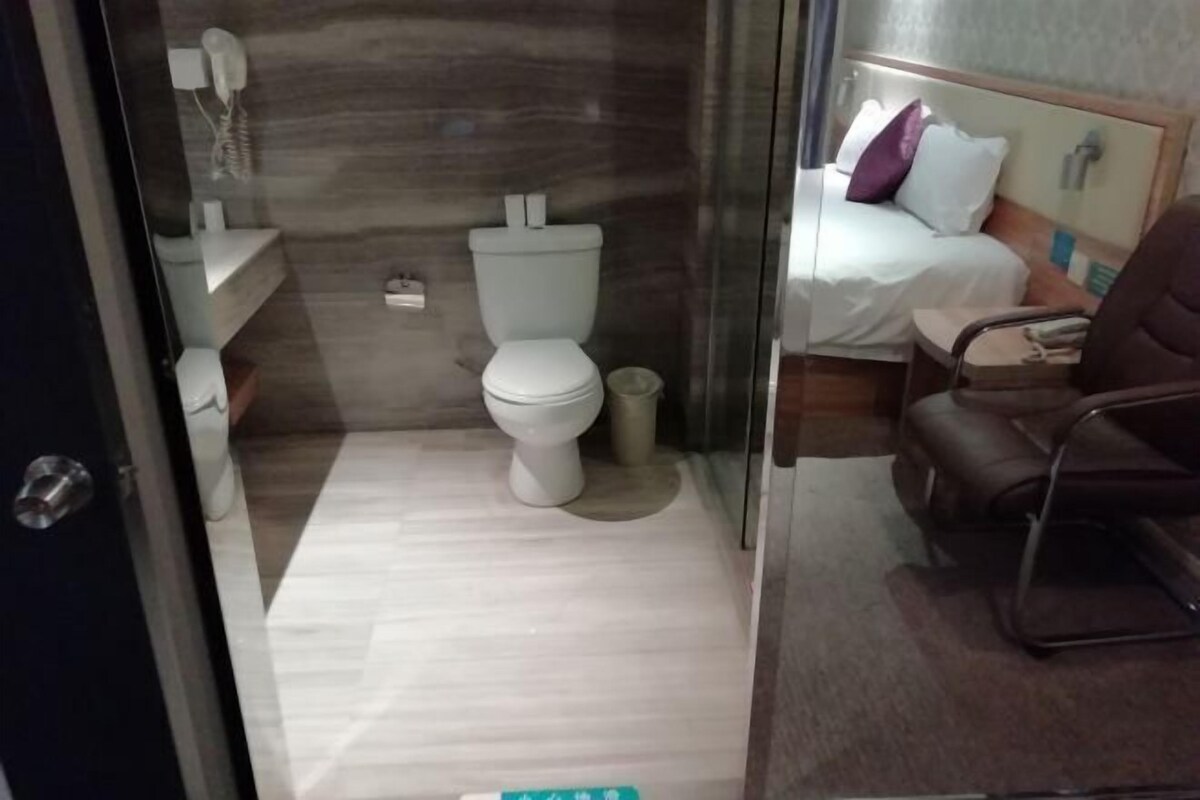 A bathroom with a toilet, a sink, and a mirror. There is a bed with a pillow and a chair in the adjacent room.