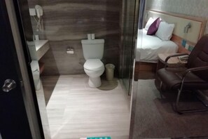 A bathroom with a toilet, a sink, and a mirror. There is a bed with a pillow and a chair in the adjacent room. - Lejia Hotel (Bazhou Shengfang International Furniture Expo City) (Bazhou)