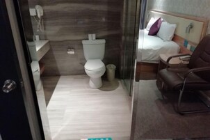 Un baño con inodoro, lavamanos y espejo. En la habitación contigua hay una cama con almohada y una silla.