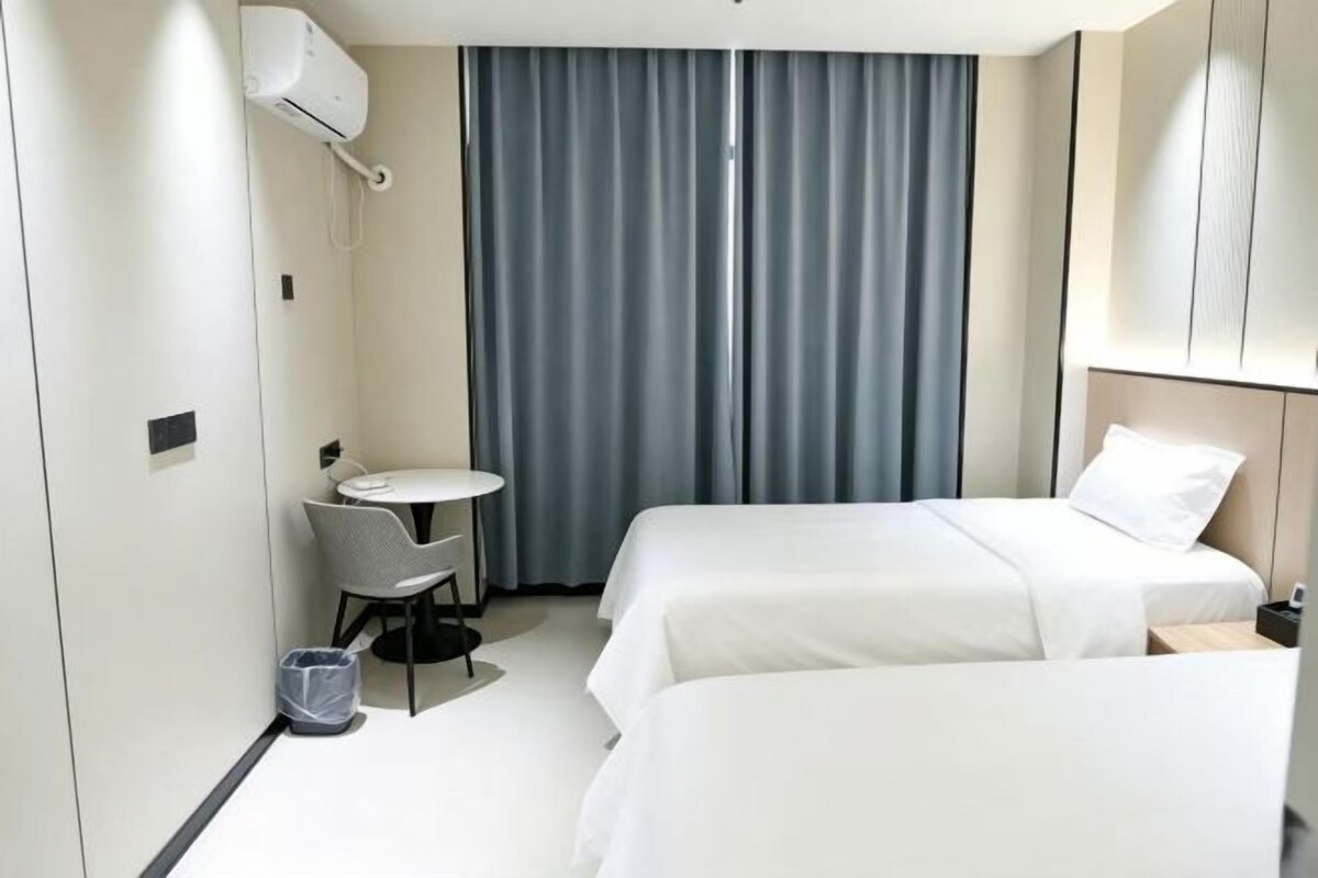Habitación de hotel con dos camas, una mesita, una silla y un ventanal con cortinas.