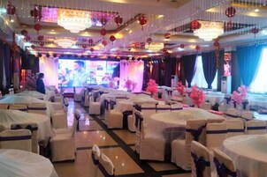 Un salón de banquetes con mesas redondas cubiertas con manteles blancos y sillas, un escenario con una pantalla grande y faroles rojos colgados del techo.