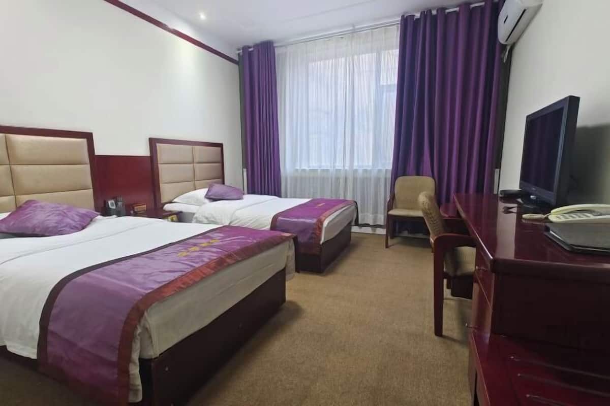 A hotel room with two beds, a desk, a chair, a television, and a window with purple curtains.