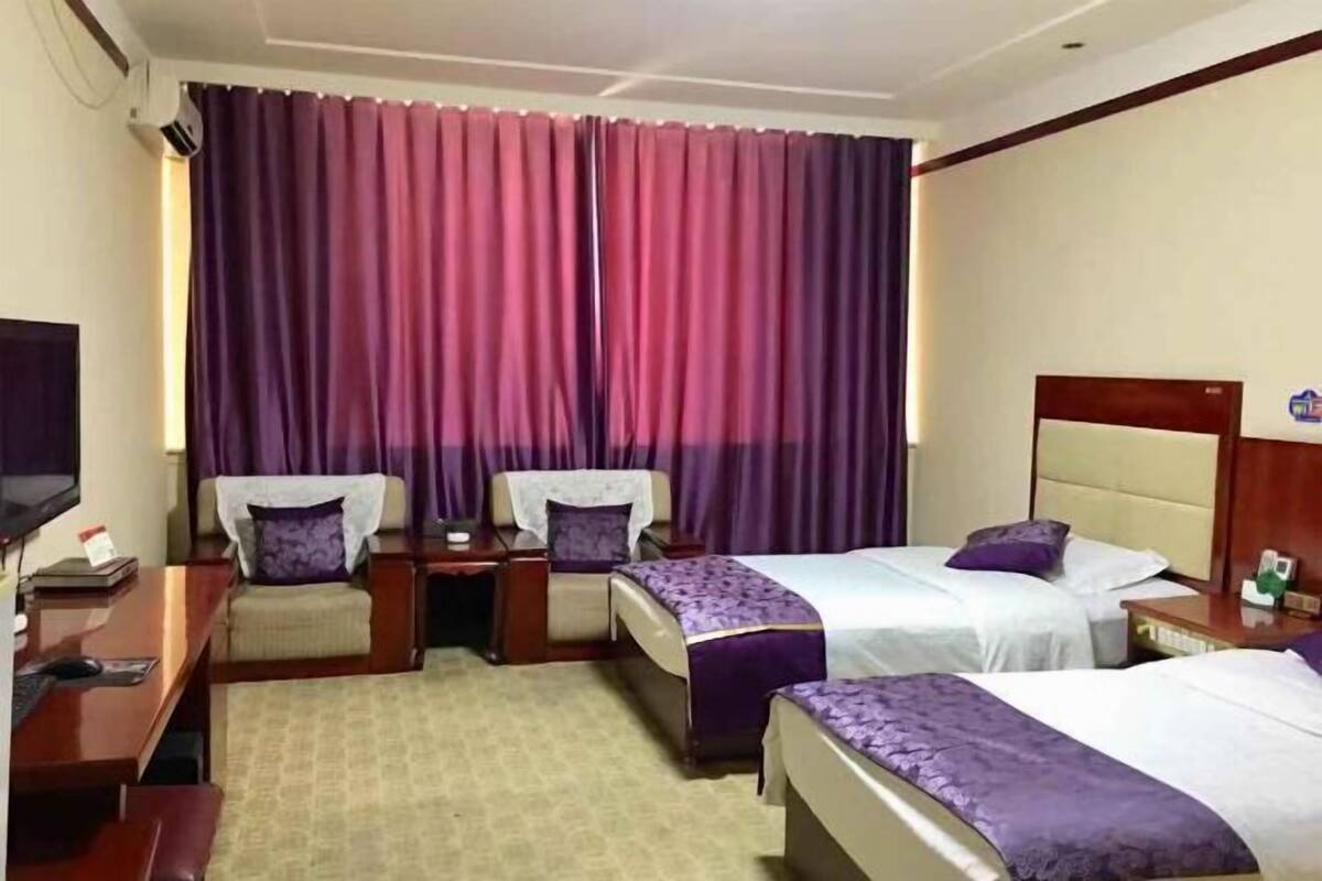 A hotel room with two beds, a desk, a chair, a TV, and a large window with purple curtains.