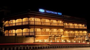 Ett hotell med en skylt som visar texten ”Mentokling Resi” och ”Mahindra”. Hotellet har en traditionell arkitektur med flera balkonger och en väl upplyst fasad.