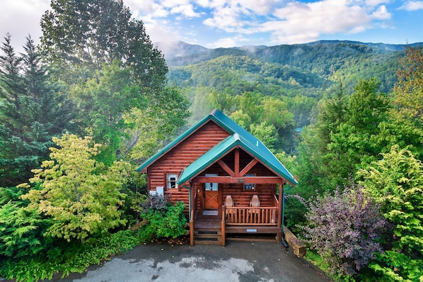 Un chalet rustique doté d’une véranda, entouré d’une végétation luxuriante et avec des montagnes en toile de fond.
