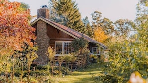 Une cabane rustique en rondins de bois, dotée d’une cheminée en pierre, entourée d’un jardin où poussent diverses plantes et arbres.