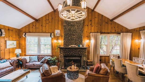 L’intérieur d’un chalet confortable, avec une cheminée en pierre, des murs en bois, un luminaire rond au plafond et un coin repas comprenant des chaises et une table.