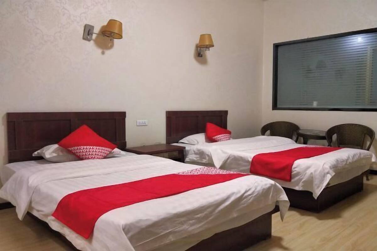 A hotel room with two beds, each with a red pillow and blanket, a wooden headboard, a small table, two wall-mounted lamps, and a window with blinds.
