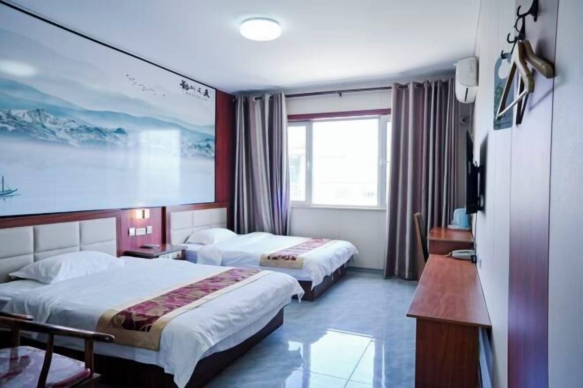 A hotel room with two beds, a desk, a chair, a window with curtains, a wall-mounted TV, and a painting.