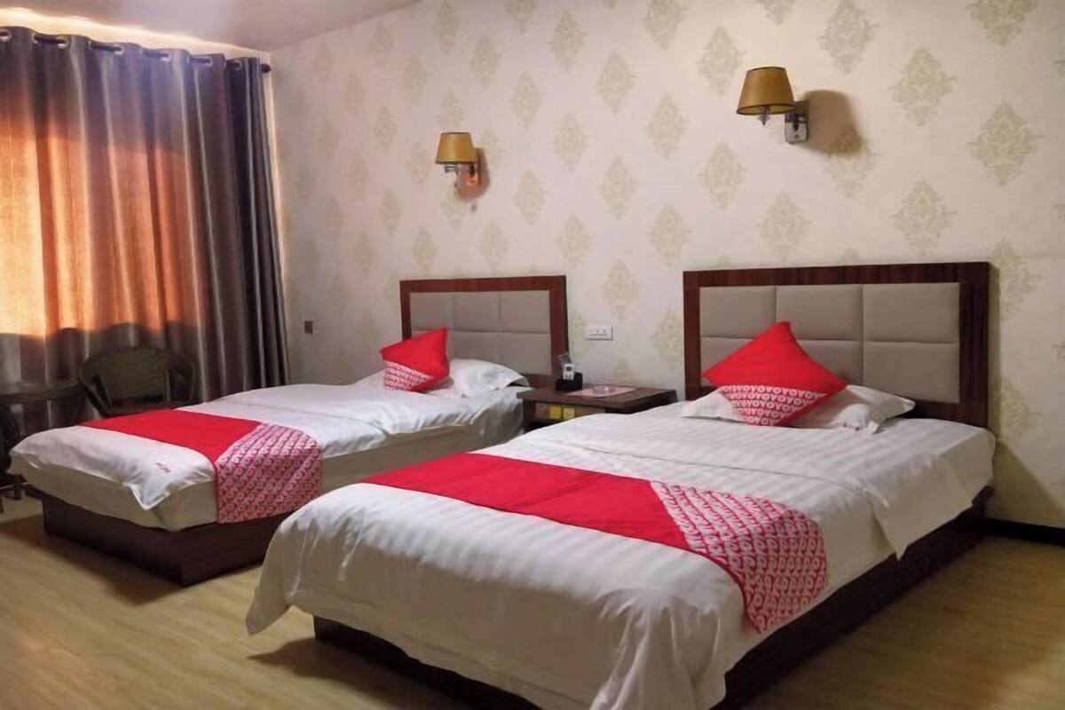 Two double beds with red pillows, a wooden headboard, a small table, a chair, and a wall-mounted lamp.