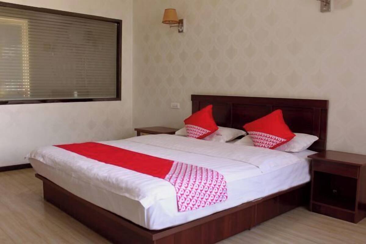 A neatly made bed with a red and white patterned bedspread, two red pillows, a wooden headboard, a bedside table, and a window with blinds.