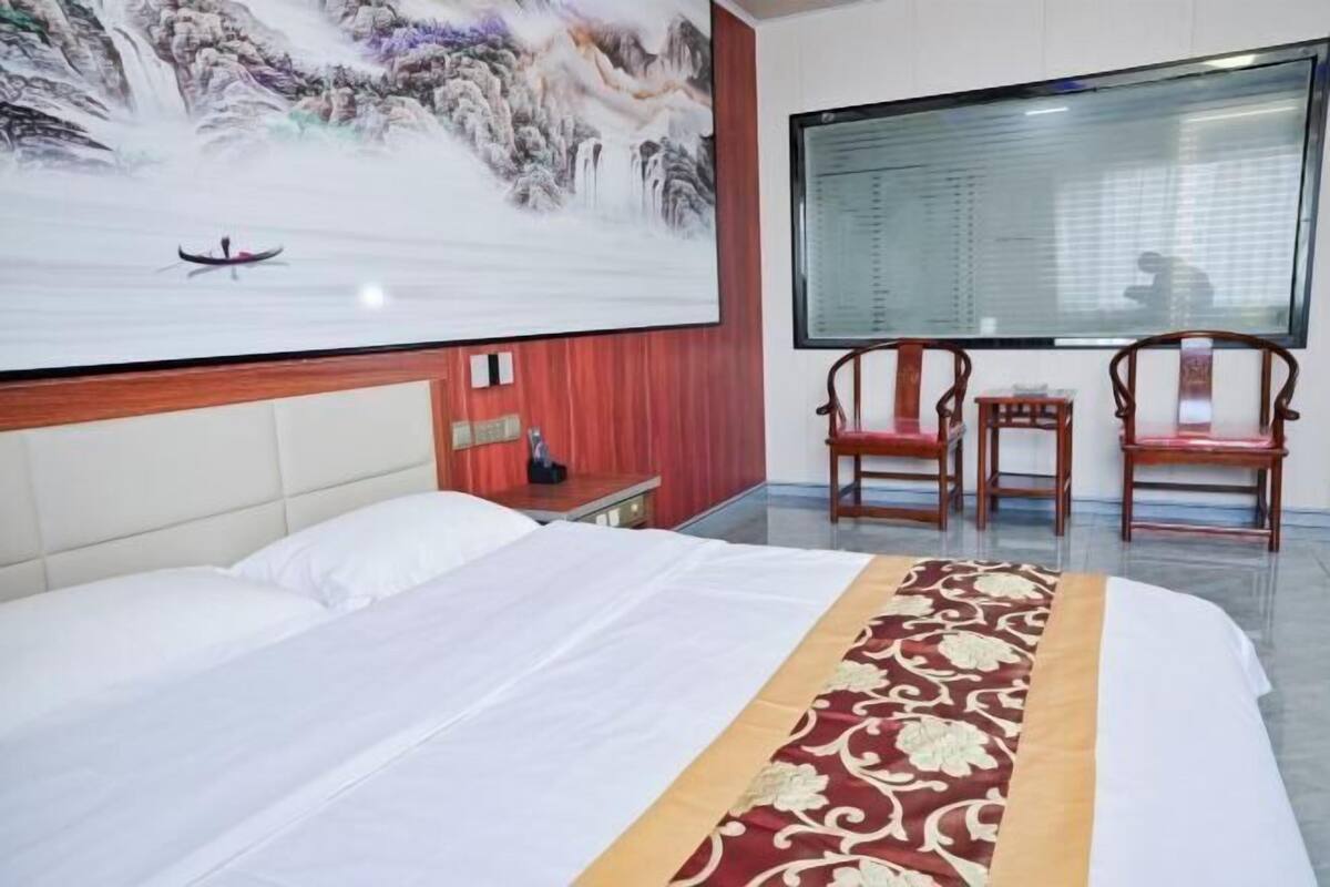 A hotel room with a bed, a painting of a mountainous landscape, two wooden chairs, and a large window.