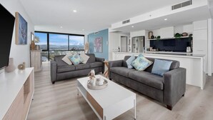 A modern living room with two grey sofas, a white coffee table, and a large TV. The room has a large window offering a city view.