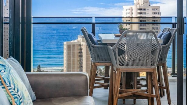 A balcony with a view of the ocean and buildings, furnished with a sofa and chairs.