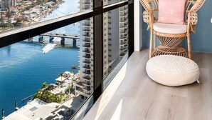 A balcony with a wicker chair and ottoman, overlooking a cityscape and a river.