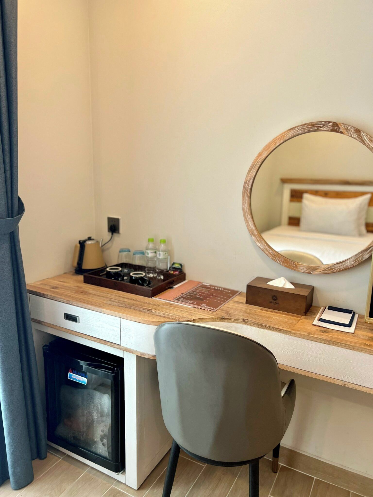 A compact hotel room with a wooden desk, a grey chair, a compact dishwasher, a bedside table with a tissue box, a mirror, and a bed with white bedding.