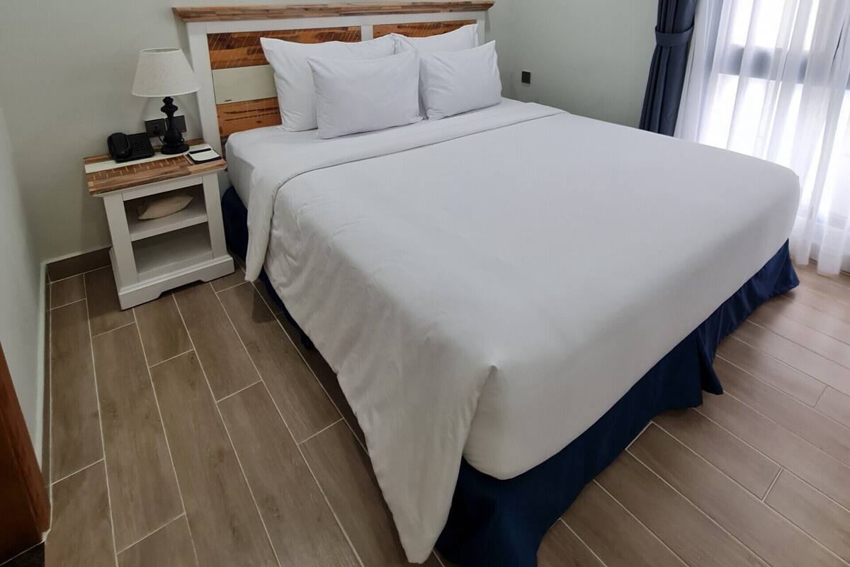 Ein ordentlich eingerichtetes Hotelzimmer mit einem großen Bett, einem Nachttisch mit Lampe und einem Fenster mit Vorhängen.