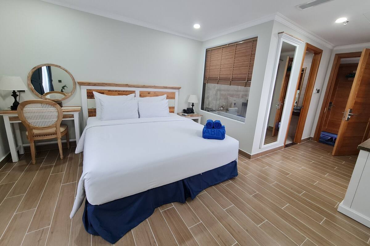 Ein Hotelzimmer mit einem großen Bett, einem Schreibtisch mit Stuhl, einem Spiegel und einem Fenster mit Jalousien.