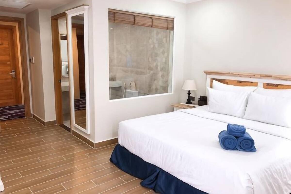 Ein Hotelzimmer mit einem großen Bett, einem Holzkopfteil, einem Fenster mit Jalousien, einem Nachttisch mit Lampe und einem durchgehenden Durchgang sichtbarem Badezimmer.