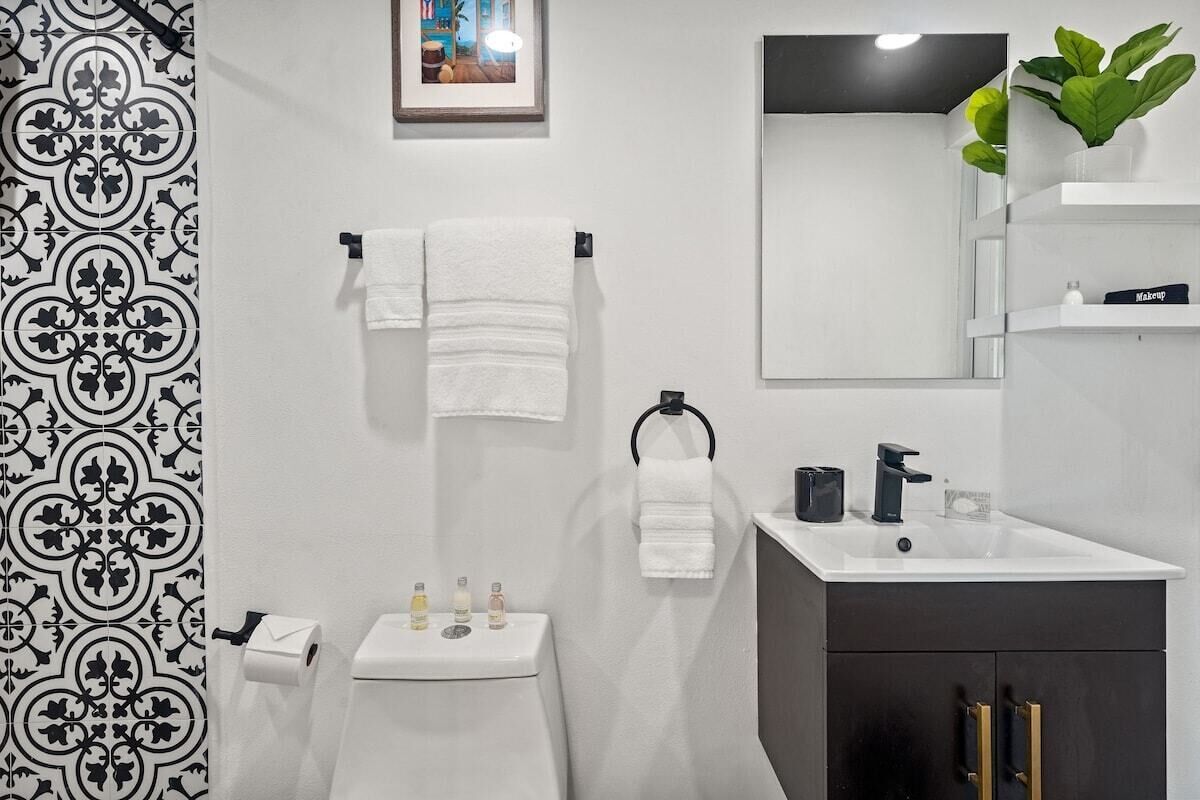Bany modern amb vàter blanc, un lavabo i un mirall. Hi ha tovalloles penjades en un suport i una planta en maceta sobre una prestatgeria.