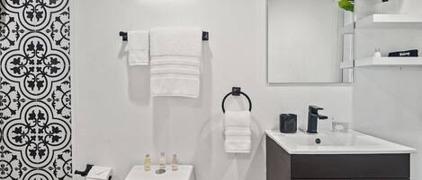 Bany modern amb vàter blanc, un lavabo i un mirall. Hi ha tovalloles penjades en un suport i una planta en maceta sobre una prestatgeria.