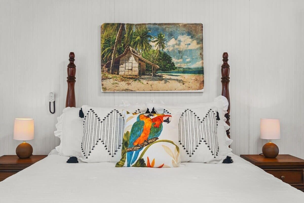 Een bed met erboven een schilderij van een tropisch strand, een wit kussen met een vogelprint in kleur, en twee bedlampen op houten nachtkastjes.