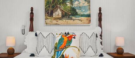 Een bed met erboven een schilderij van een tropisch strand, een wit kussen met een vogelprint in kleur, en twee bedlampen op houten nachtkastjes.