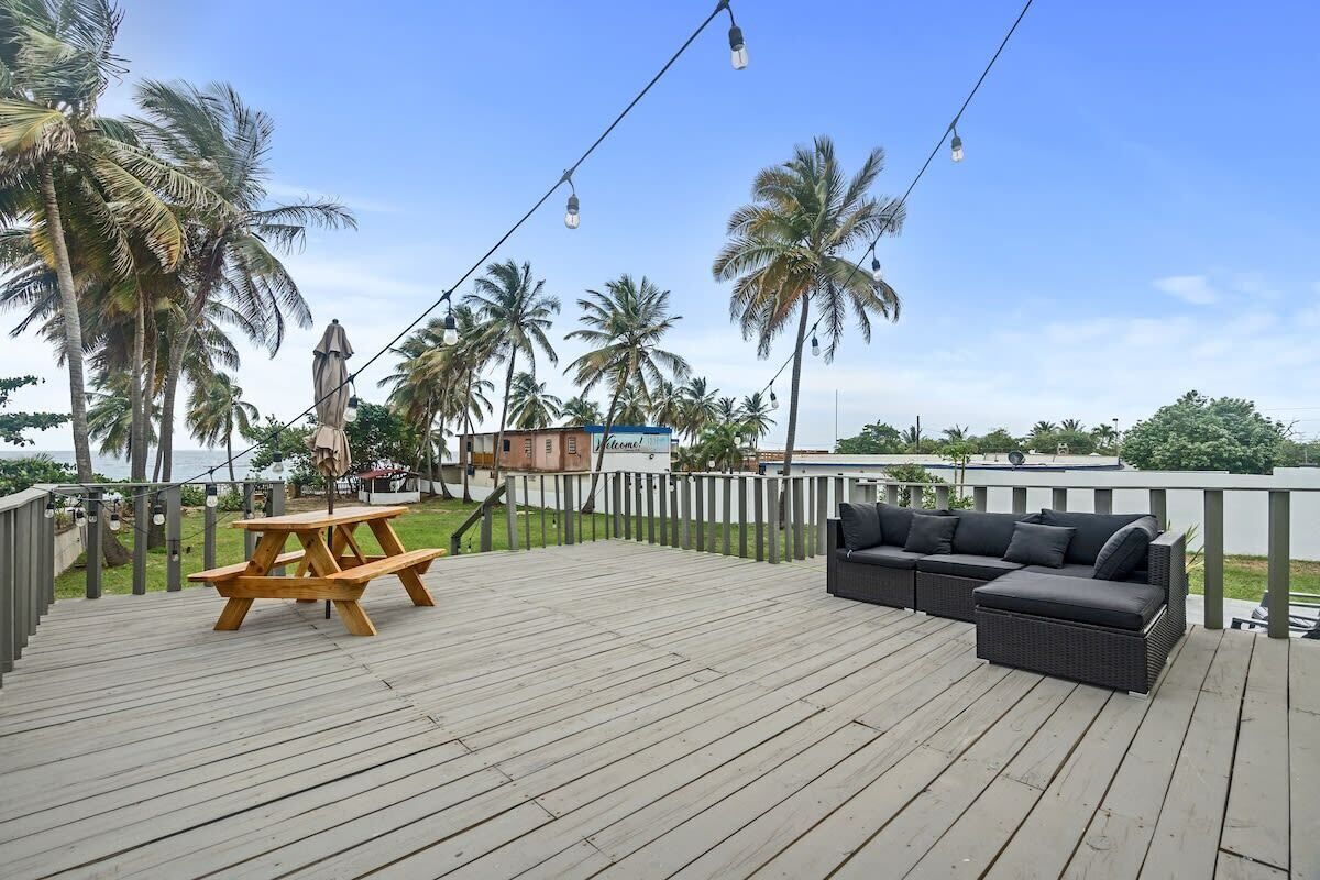 A wooden deck with a picnic table and a sofa, surrounded by palm trees and a view of the ocean.