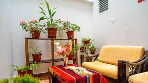 Un espace de détente intérieur, chaleureux, avec une chaise en bois et une petite table recouverte d’un tissu coloré. On y trouve plusieurs plantes en pot, disposées sur des étagères, ainsi qu’un banc en bois.