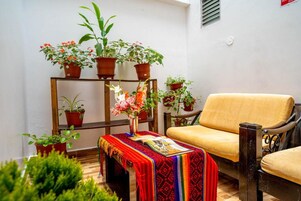 Un rincón interior acogedor con una silla de madera y una mesa pequeña cubierta con un paño colorido. Hay varias plantas en macetas sobre estantes y un banco de madera.