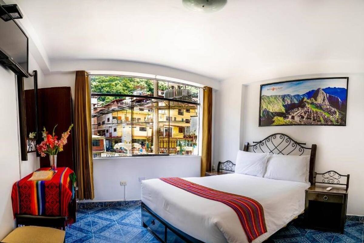 Een hotelkamer met een bed, een nachtkastje, een aan de muur bevestigde televisie, een raam met uitzicht op gebouwen en een schilderij.