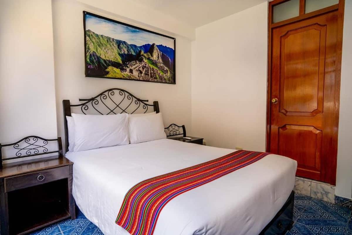Een keurig opgemaakt bed met een kleurrijke gestreepte deken, een houten deur en een schilderij van een bergachtig landschap aan de muur.
