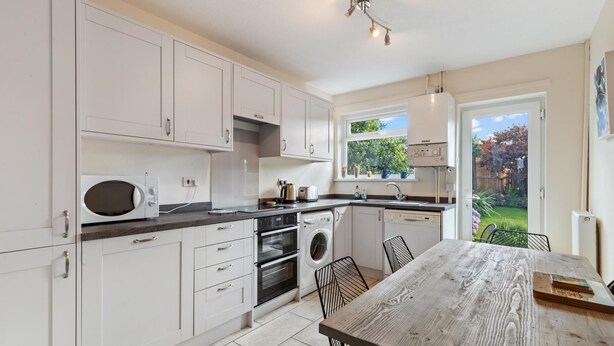 A modern kitchen with white cabinets, a wooden dining table, and a view of a garden through the window. - Ballyroan - Peaceful Dog Friendly Cottage (Tenby)