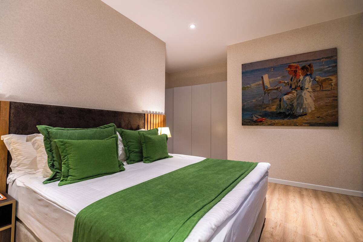 Ett hotellrum med en stor säng, gröna kuddar, en tavla på väggen och ett trägolv.