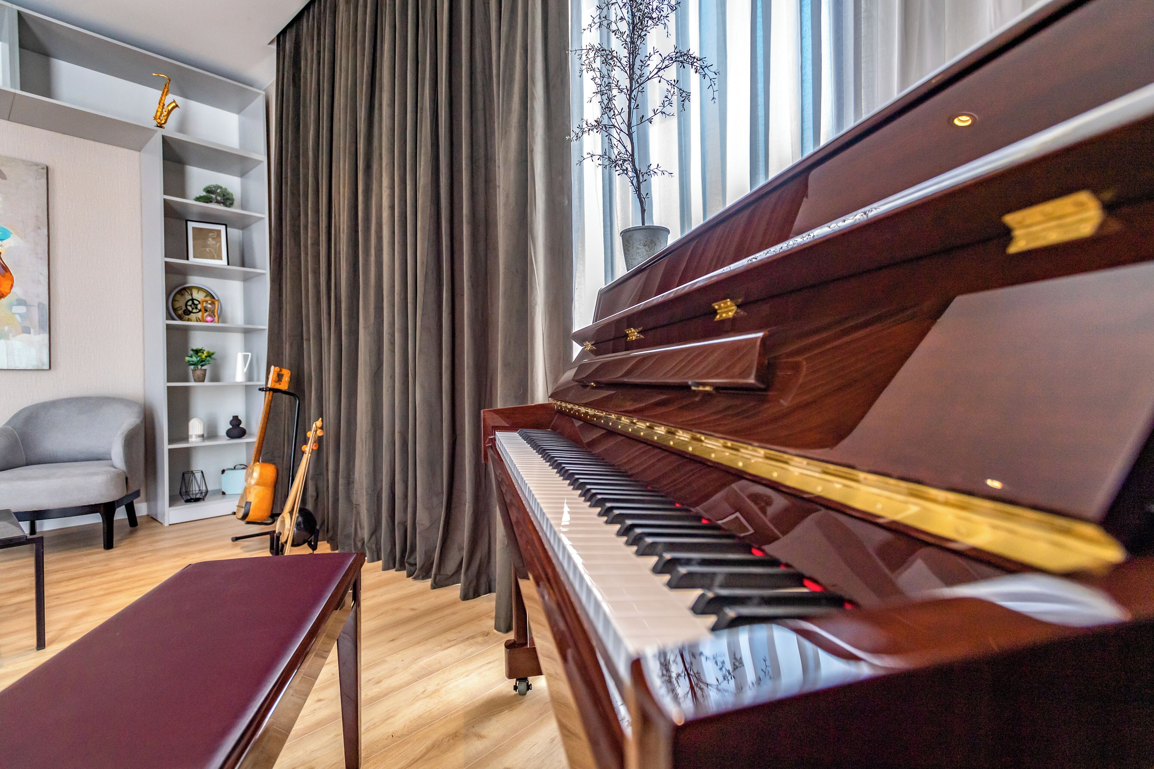 A grand piano in a well-lit room with a wooden floor, a grey armchair, and a bookshelf with various items.