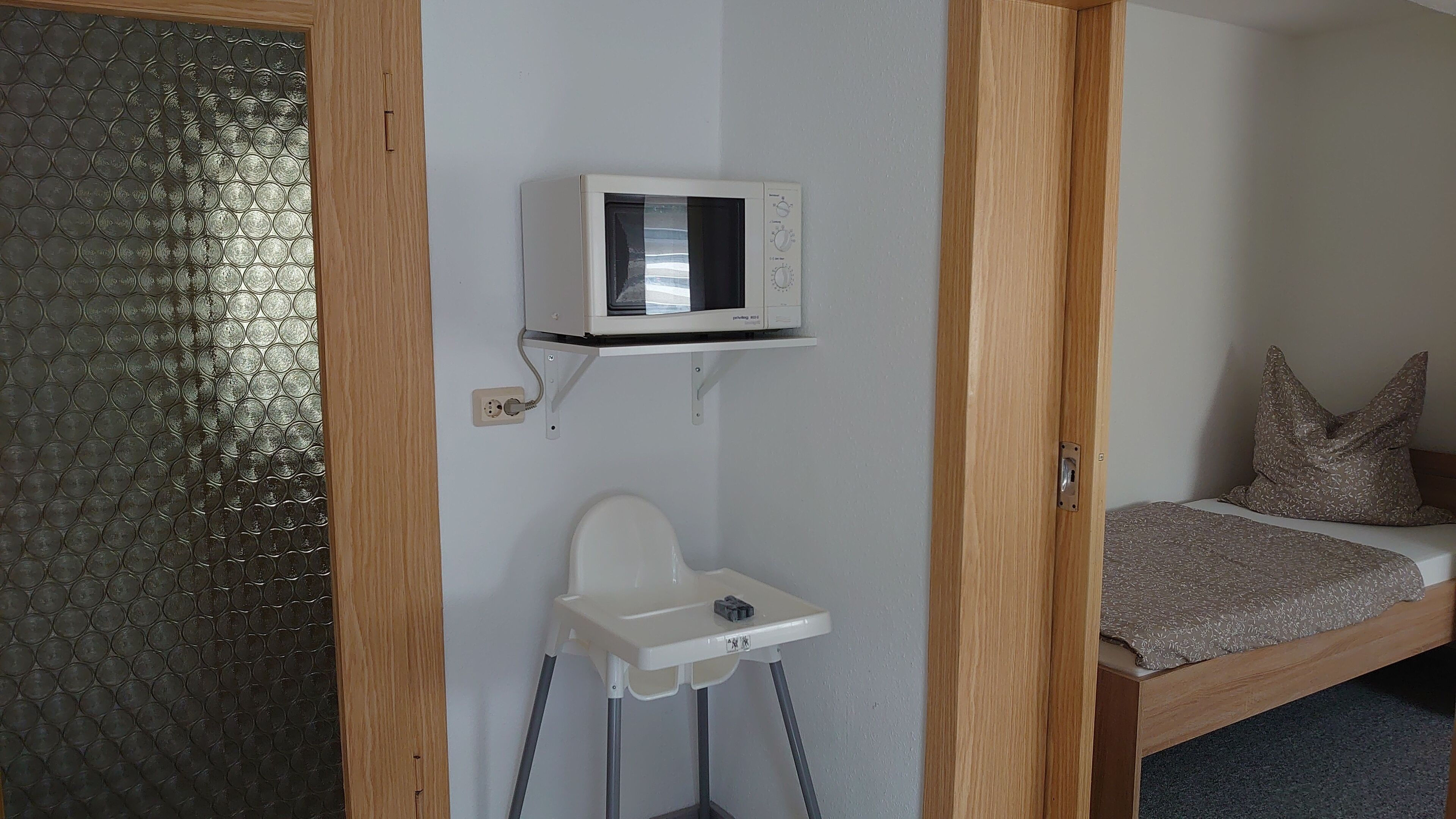 A compact room with a microwave mounted on the wall, a small white chair, and a bed with a patterned blanket.