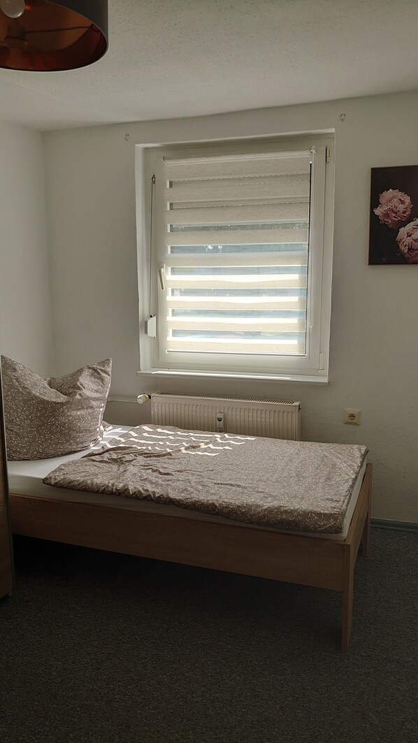 Een slaapkamer met een enkel bed, een radiator, een raam met zonwering en een wandcontactdoos.