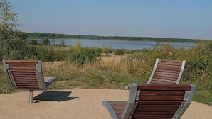 Two wooden benches with metal frames on a gravel surface, overlooking a lake and distant trees.
