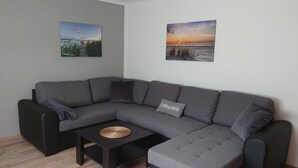A grey sectional sofa with a woven coffee table in front, two wall-mounted pictures depicting landscapes, and a small grey pillow on the sofa.