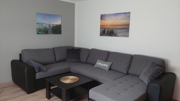 Um sofá modular cinza com uma mesa de centro trançada na frente, duas quadros fixados na parede retratando paisagens, e uma pequena almofada cinza no sofá.