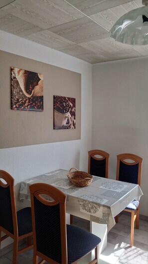 Uma sala de jantar com uma mesa posta para quatro pessoas, cadeiras de madeira e dois quadros pendurados na parede.