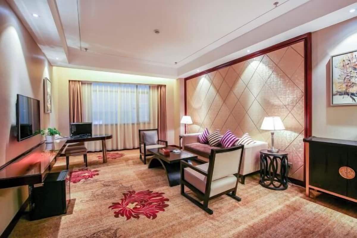 A hotel room with a TV, sofa, chairs, a small table, a lamp, and a decorative wall panel.