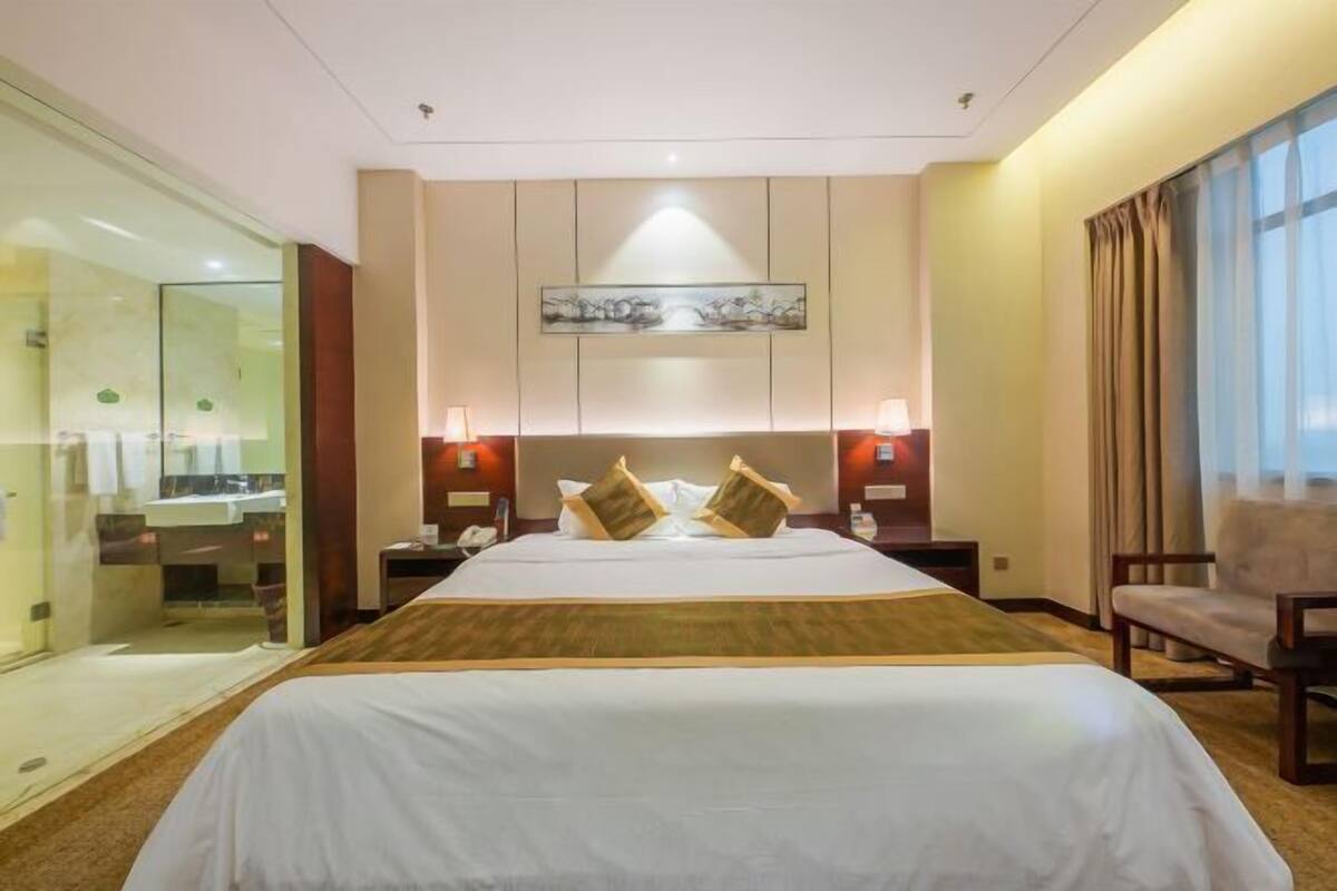 A hotel room with a large bed, bedside tables, a mirror, a window with curtains, and a wall-mounted artwork.