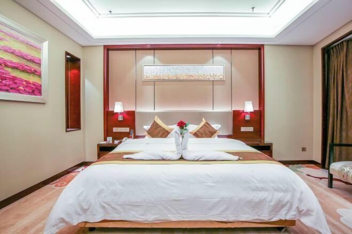 A hotel room with a large bed, two bedside tables, a nightstand, a wall-mounted artwork, and a ceiling light.