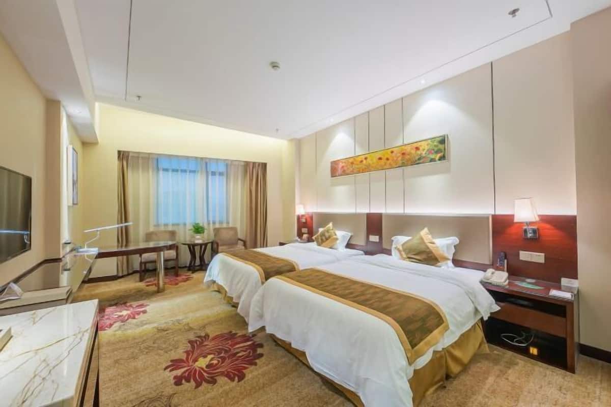 A hotel room with two beds, a desk, a TV, and a large window with curtains.