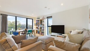A modern living room with a large flat-screen TV, a bookshelf, and a balcony with outdoor seating. - Lovely Luminous 3 Bed Flat, With Secure Ev Parkng (London)
